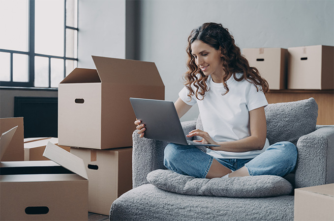 A woman on a laptop sitting on a chair surrounded by moving boxes.